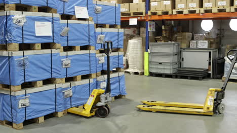 Warehouse-worker-in-high-visibility-vest-walks-between-rows-of-blue-wrapped-pallets,-using-a-Hand-Pallet-Truck-with-Double-Rollers