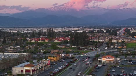 Zeitraffer-über-Einer-Belebten-Kreuzung-In-San-Bernardino,-Kalifornien,-Bei-Sonnenuntergang-Mit-Rosa-Wolken-Im-Hintergrund-Vor-Den-Schneebedeckten-Bergen-Des-San-Bernardino-National-Forest,-Luftbild