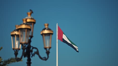 La-Bandera-De-Los-Emiratos-Árabes-Unidos-Ondeando-En-El-Asta-De-La-Bandera,-Enmarcada-Por-Un-Poste-De-Luz-Tradicional,-En-La-Isla-De-La-Bandera-De-Sharjah-En-Los-Emiratos-Árabes-Unidos.