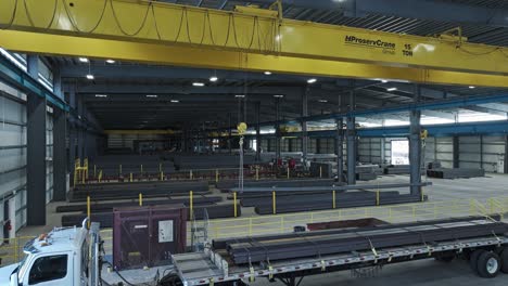 Yellow-overhead-bridge-crane-in-warehouse-moves-across-tracks-carrying-steel-beam-in-warehouse