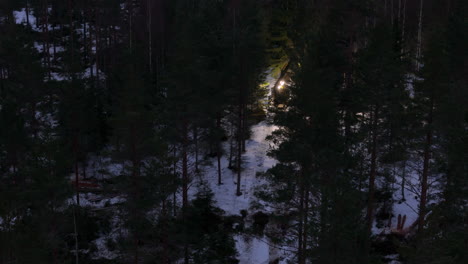 Telephoto-night-drone-shot-of-tree-harvester-navigating-snowy-track-in-forest