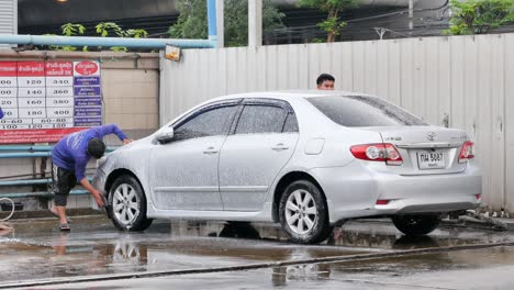 Drei-Männer-Wischen-Die-Karosserie-Eines-Autos-Ab-Und-Helfen-Sich-Gegenseitig-Beim-Reinigen-Mit-Wasser-Und-Seife-In-Einer-Autowaschanlage-In-Bangkok,-Thailand