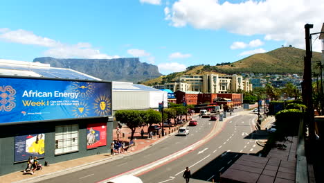 Cars-and-pedestrians-walk-at-touristic-Cape-Town-Waterfront-with-views-of-iconic-Table-Mountain-and-Signal-Hill-against-blue-sky
