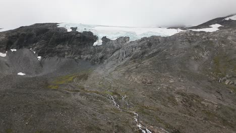 Ein-Großer-Gletscher-Auf-Einem-Felsigen-Berg-Mit-Einigen-Kleinen-Wasserfällen-Und-Flüssen-In-Einer-Nebligen-Umgebung,-Norwegen,-Europa,-Drohne