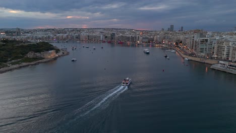An-aerial-view-of-the-Malta-Sliema-to-Valletta-ferry-making-its-way-into-Sliema-harbour-at-dusk,-with-the-resort-town-in-the-background
