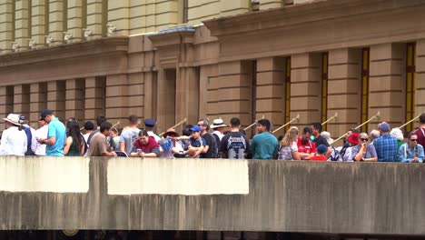 Australian-families-lined-up-on-the-sky-bridge-above-Adelaide-street,-waiting-for-the-commencement-of-annual-Anzac-day-parade-tradition,-watching-the-marching-parade