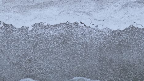 Borde-De-Un-Lago-Congelado-Con-Hielo-Que-Se-Encuentra-Con-Una-Orilla-Cubierta-De-Nieve,-A-Vista-De-Pájaro,-A-La-Luz-Del-Día