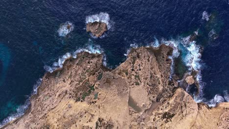 Vista-De-Arriba-Hacia-Abajo-Sobre-Una-Costa-Rocosa-En-La-Bahía-De-Dwejra,-Con-Olas-Chapoteando