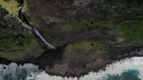 Madeira-Véu-da-Noiva-viewpoint-aerial-view-descending-rugged-cliff-waterfall-cascading-into-Atlantic-ocean-shore