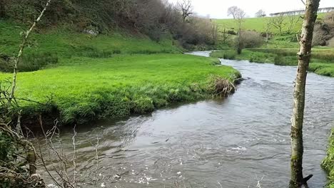 Curving-flooded-river-running-through-Welsh-valley-farmland-meadow-slow-motion