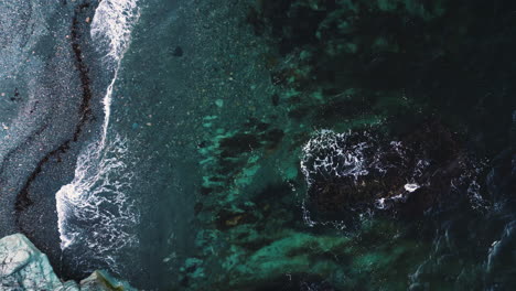 Ocean-waves-swirl-in-shallow-grey-beach-with-pronounced-tide-line-of-seaweed,-drone-top-down