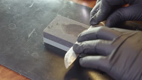 Hands-in-black-gloves-sharpen-kitchen-knife-on-wet-whetstone-on-table