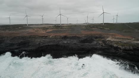 Aerial-panning-shot-of-a-wind-farm-at-background-with-ocean-water-hitting-the-coast-of-Cape-Bridgewater,-Australia