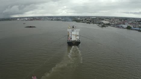 Cargo-ship-sailing-in-the-Suriname-river-towards-harbor-with-ferry-boat,-drone-view,-city-in-background