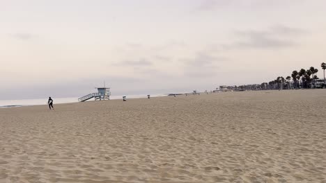 Surfer-With-Surfboard-Running-On-Sandy-Shore-Towards-Sea-In-Venice-Beach,-California