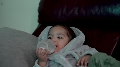 British-Asian-Toddler-Wearing-Bathrobe-Laying-On-Sofa-Drinking-Milk-From-Bottle-With-Cheeky-Expression-On-Face