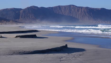 Sand-blows-off-Atacama-Desert-mountains-over-ocean-beach-waves,-Chile
