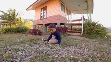 Toddler-playing-with-stones-sitting-on-the-ground-,-in-front-of-a-small-farm-house-loated-at-the-hill-top-plantation-in-Phetachabun-Thailand