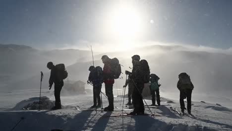Group-of-people-standing-in-snowy-mountain-during-windy-hike,-silhouette-in-front-of-sun,-Slow-motion