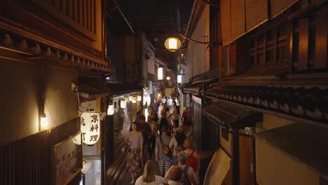 Toma-Nocturna-En-Movimiento-A-Través-Del-Abarrotado-Callejón-Pontocho,-Shijo-dori,-Con-Exteriores-Tradicionales-De-Madera-Y-Linternas-Que-Iluminan-El-Distrito-De-Nakagyo,-Lleno-De-Turistas,-Kyoto,-Japón-1