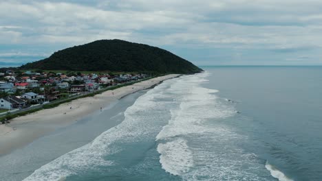 Rising-Aerial-of-Ocean-Waves-Hitting-Beach-Near-Coastal-Town,-Hill-in-Background