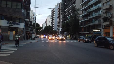 Busse,-Taxis-Und-Verkehr-In-Der-Stadt-Buenos-Aires.-Fußgänger-Gehen-Auf-Der-Corrientes-Avenue,-Sonnenuntergangs-Skyline-Zwischen-Großstadtgebäuden-Und-Geschäften