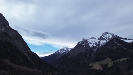 Drone-Moviéndose-Lentamente-Sobre-Un-Bosque-De-Pinos-Con-Cumbres-Cubiertas-De-Nieve-En-El-Fondo-En-Vorarlberg,-Austria
