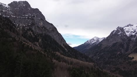 Drone-rise-up-hill-and-fly-towards-pine-forest-surrounded-by-snow-covered-summits