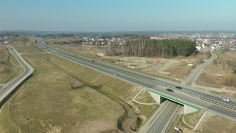 Aerial-shot-of-a-highway-intersection-with-overpass,-vehicles-in-transit,-and-surrounding-early-spring-landscape