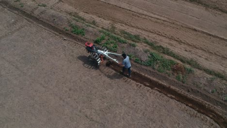 Shooting-from-drone-flying-over-tractor-with-harrow-system-plowing-ground-on-cultivated-farm-field,-pillar-of-dust-trails-behind,-preparing-soil-for-planting-new-crop,-agriculture-concept,-top-view