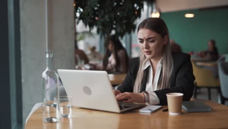 beautiful,-sad-young-woman-sits-in-a-cafe-in-business-attire,-saddened-by-what-she-sees-on-her-laptop,-grasping-her-head