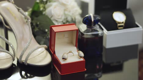 A-red-box-with-earrings-and-a-wedding-ring-next-to-a-perfume-bottle-and-a-watch