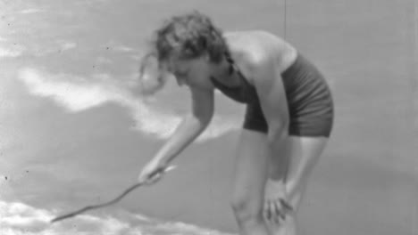 1930s-Elegant-Woman-in-Swimsuit-Writing-in-Sand-at-Beach