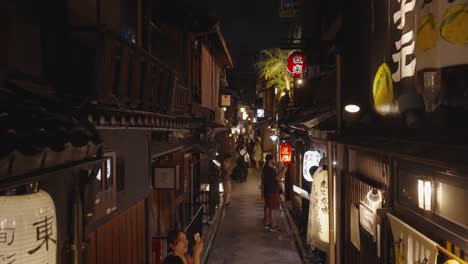 Toma-Nocturna-En-Movimiento-A-Través-Del-Abarrotado-Callejón-Pontocho,-Shijo-dori,-Con-Exteriores-Tradicionales-De-Madera-Y-Linternas-Que-Iluminan-El-Distrito-De-Nakagyo,-Lleno-De-Turistas,-Kyoto,-Japón-2