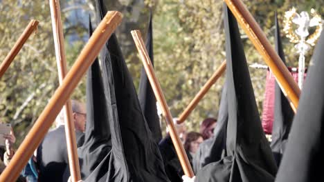 Back-view-penitent-marching-on-black-tunic-with-hoods-holding-long-brown-candles-across-and-red-banner-on-silver-stand