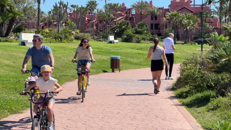 People-walking-and-riding-bicycles-on-a-sunny-day-near-Estepona-beach-sea-side,-palm-trees-and-blue-sky,-fun-holiday-vacation-in-Spain,-Paseo-Maritimo,-4K-shot