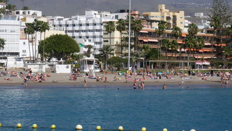 Playa-De-Los-Christianos:-Animada-Escena-De-Playa-Con-Bañistas-Y-Bañistas,-Enmarcada-Por-Palmeras-Y-Un-Telón-De-Fondo-De-Edificios-De-La-Ciudad