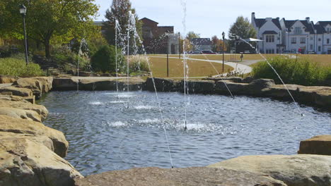 Public-park-pond-area-with-several-fountains-spraying-streams-of-clear-water-with-homes-and-buildings-in-the-background,-on-a-sunny-day