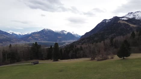 Luftaufnahme-Einer-Drohne,-Die-An-Einem-Bewölkten-Tag-In-Vorarlberg,-Österreich,-über-Hütten-Auf-Einem-Hügel-Mit-Schneebedeckten-Bergen-Im-Hintergrund-Fliegt