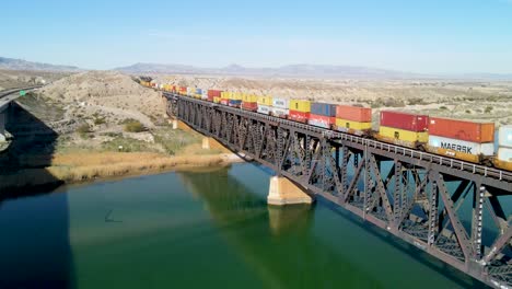 Freight-Train-Carrying-Shipping-Containers,-Multi-Color,-Red,-Yellow,-Over-Steel-Bridge-Between-California-and-Arizona-,-Colorado-River-,-I-40-freeway-East,-Wide-Angle-Aerial