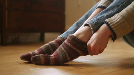 Woman-putting-on-woolen,-striped-socks-while-sitting-on-wooden-floor-in-apartment