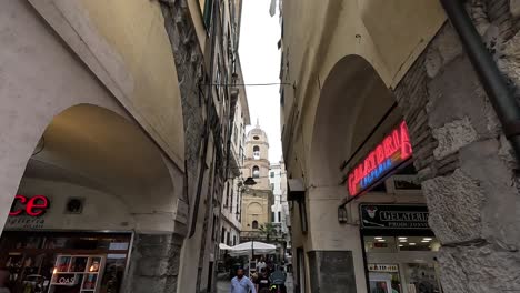 Overhead-clip-showing-narrow-city-passageway-in-Genoa,-Italy,-with-people-walking-through-and-busy-streets-with-cafes-and-restaurants