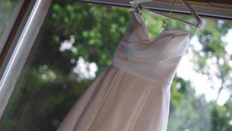 Close-up-of-bride's-white-satin-dress-hanging-on-open-window-in-room