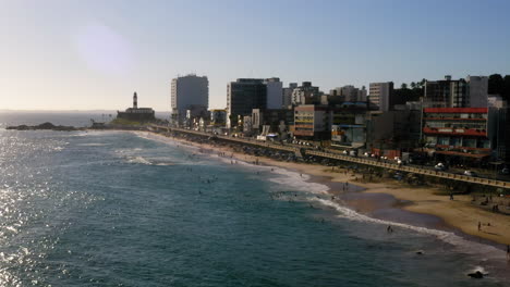 Luftaufnahme-Des-Strandes-Farol-Da-Barra-Mit-Den-Menschen,-Die-Den-Sonnigen-Tag-Genießen,-Und-Den-Gebäuden-Im-Hintergrund,-Salvador,-Bahia,-Brasilien