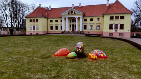 Countryside-house-with-eastern-painted-colorful-eggs-displayed-in-green-garden-grass,-european-style-home-building-in-north-eastern-europe-with-red-tiles-,-yellow-pink-pale-painted