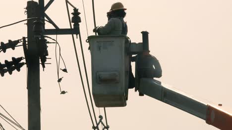 Hoisted-up-on-a-crane,-two-repairmen-are-removing-some-electrical-parts-and-installing-another-around-a-porcelain-suspension-insulator-for-a-lamppost-in-Bangkok,-Thailand