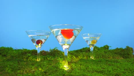 one-martini-glass-resting-on-moss-splitting-into-three-martini-glasses-with-various-fruits-being-dropped-into-the-clear-spirits-inside-including-a-strawberry,-cherry,-and-an-olive-blue-background
