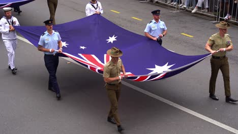 Soldaten-Der-Drei-Zweige-Der-Australischen-Streitkräfte,-Der-Royal-Australian-Navy,-Der-Australian-Army-Und-Der-Royal-Australian-Air-Force,-Marschieren-Mit-Flaggen-Bei-Der-Anzac-Day-Parade-Die-Straße-Entlang