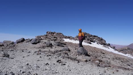 Mann-Geht-Auf-Berggipfelpfad-Vorbei-An-Schneeflecken-Vor-Blauem-Himmel