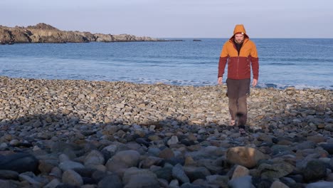 Lone-man-in-warm-clothes-walks-toward-camera-on-morning-rocky-beach
